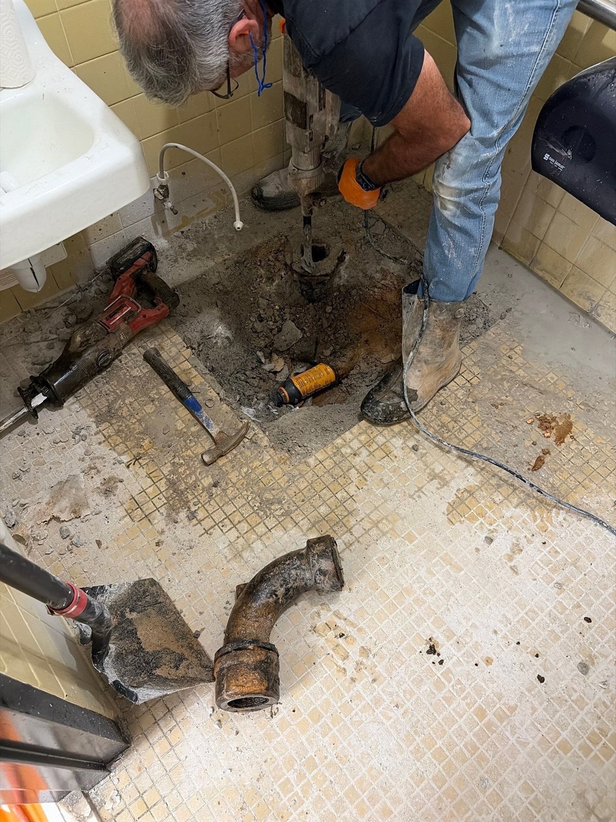 LiningWorks technician breaking out a concrete floor to reach an old cast iron drain pipe, with a removed cast iron elbow in front